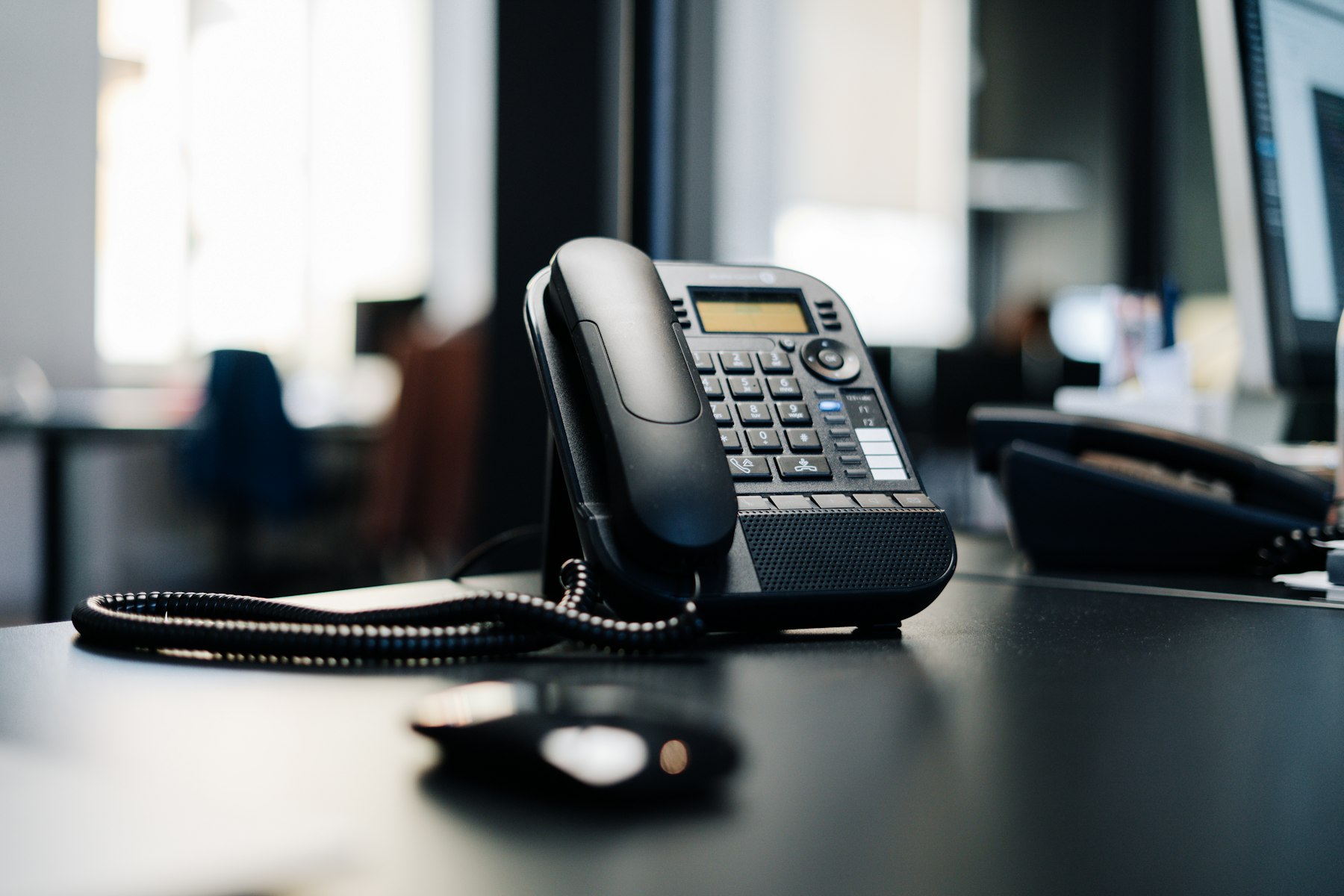 A modern office desk phone sitting on a dark desk near a computer monitor