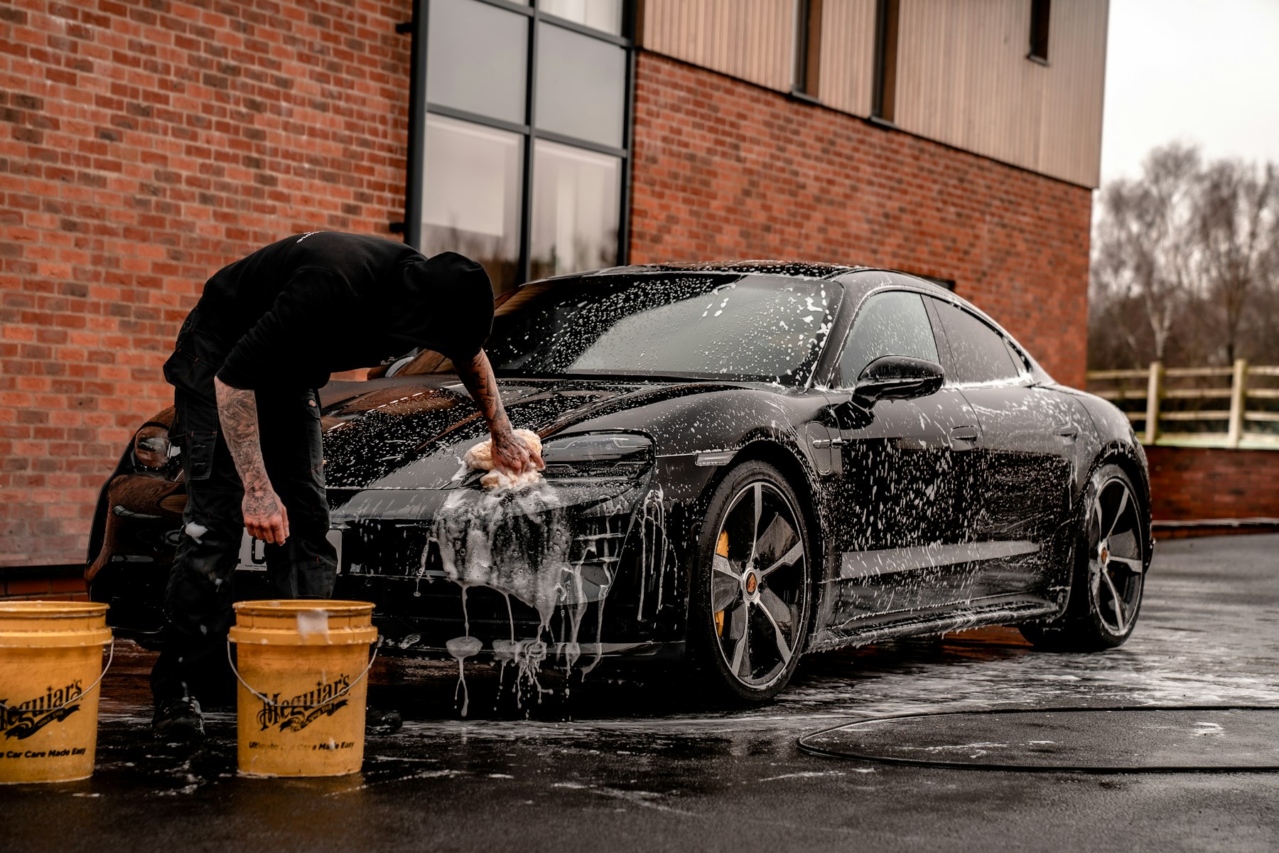 A car wash employee hand-scrubbing a black car covered in soap suds outside a brick building