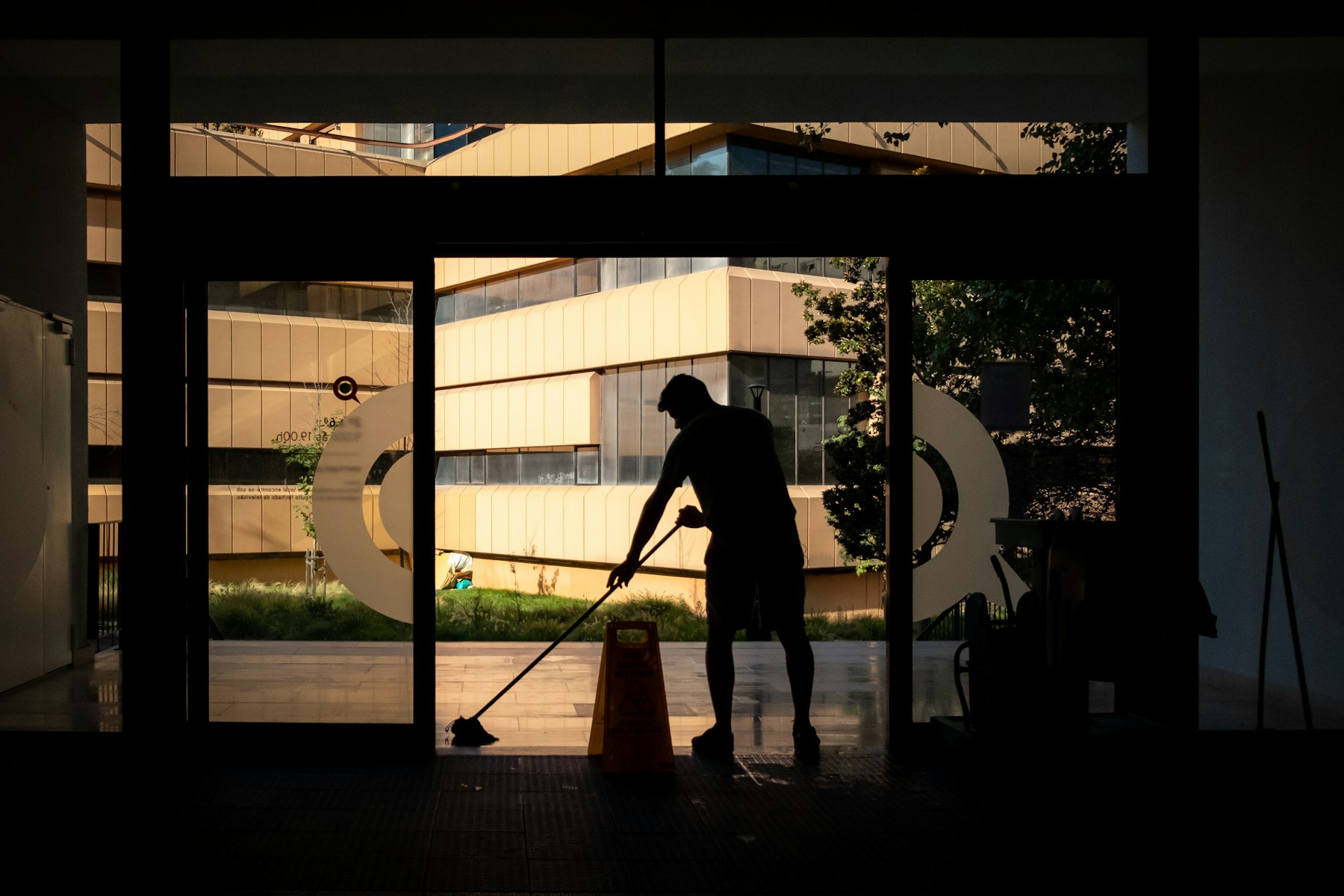 Silhouette of a janitorial worker mopping the entrance of a commercial office building at dusk