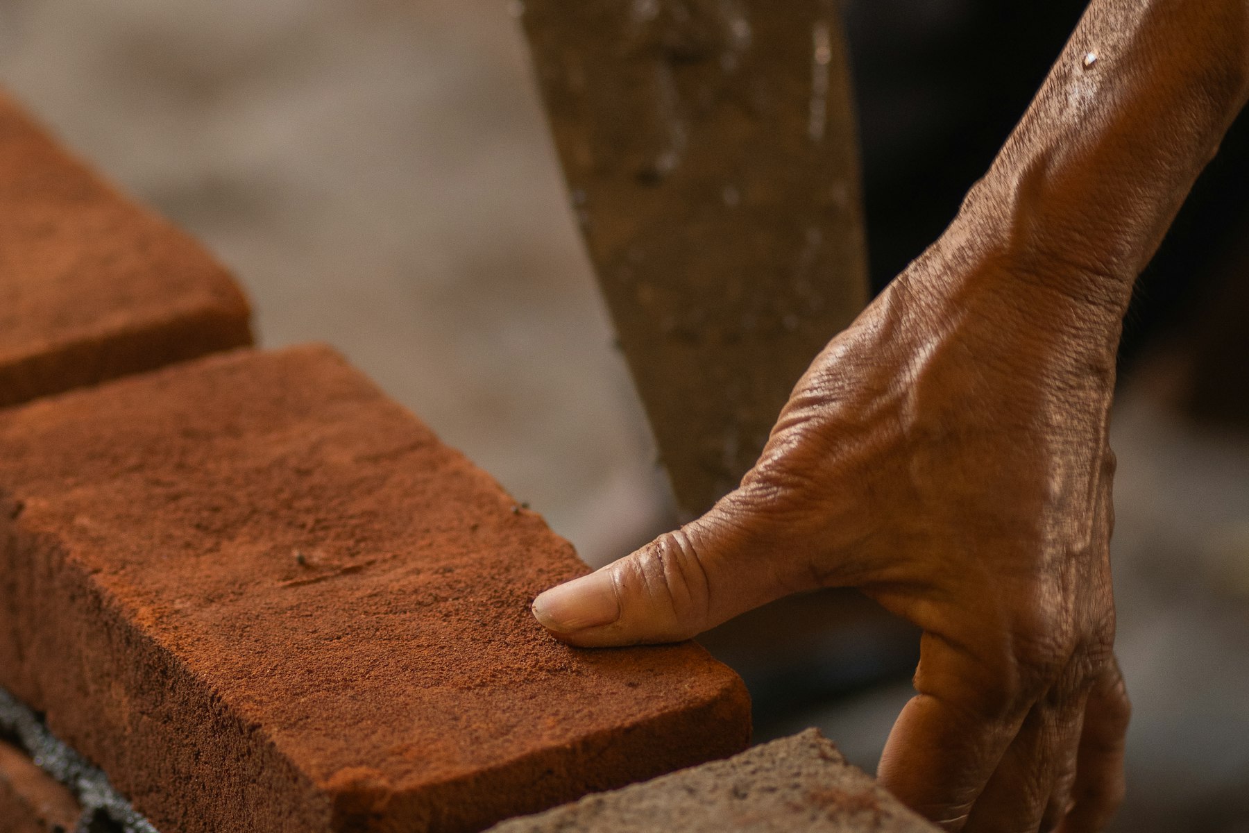 A bricklayer's hand pressing a red brick into place on a mortared wall, trowel resting nearby