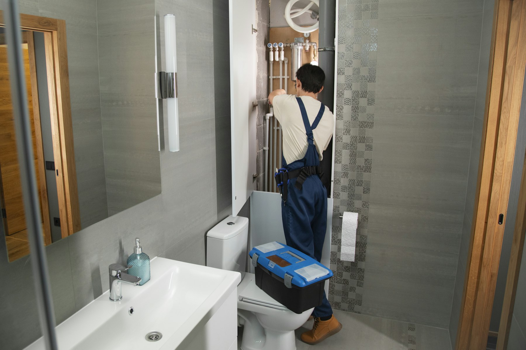 A plumber working on pipes behind a bathroom wall, toolbox nearby