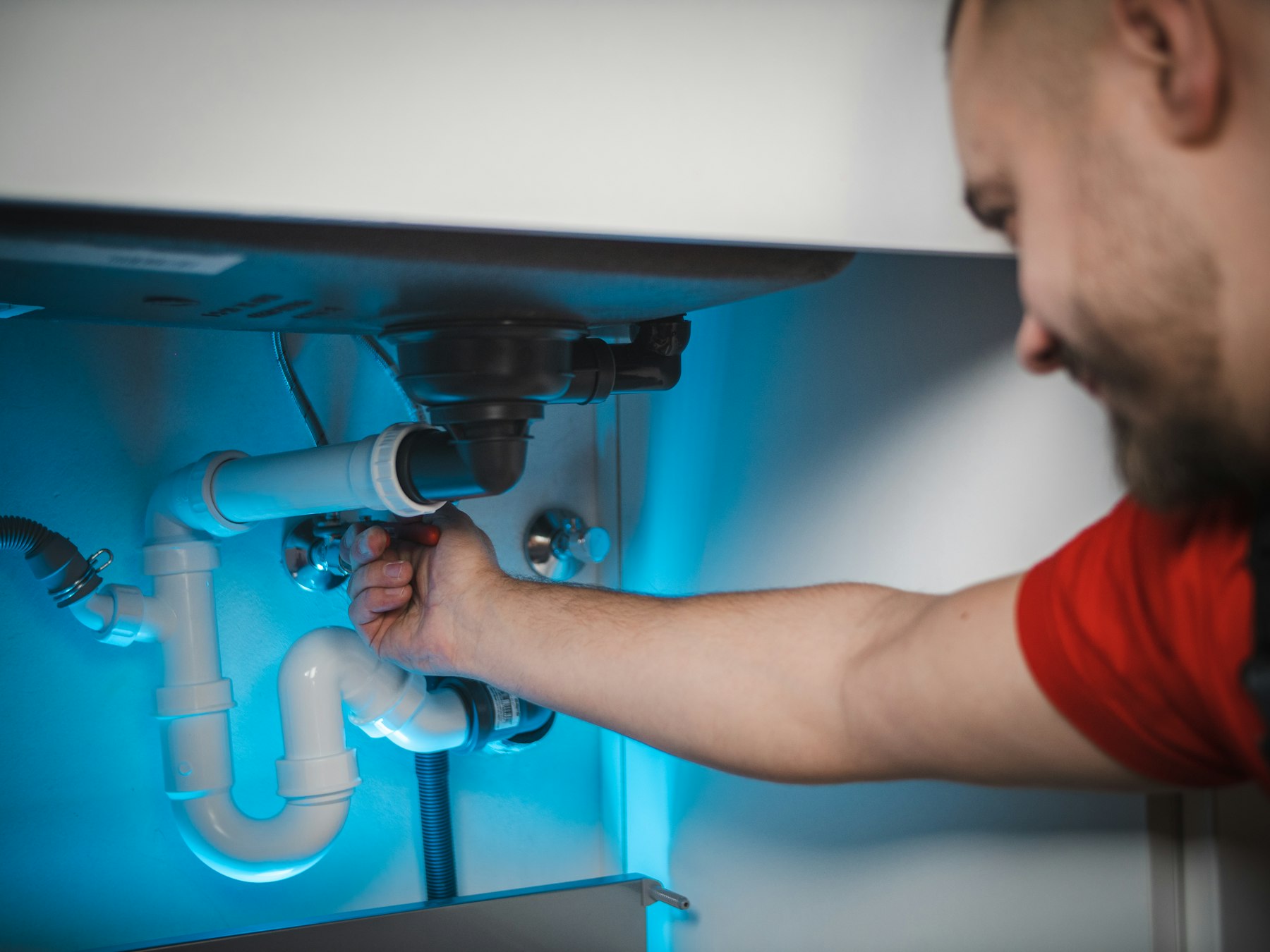 A plumber reaching under a sink to work on white PVC drain pipes, lit by a blue work light