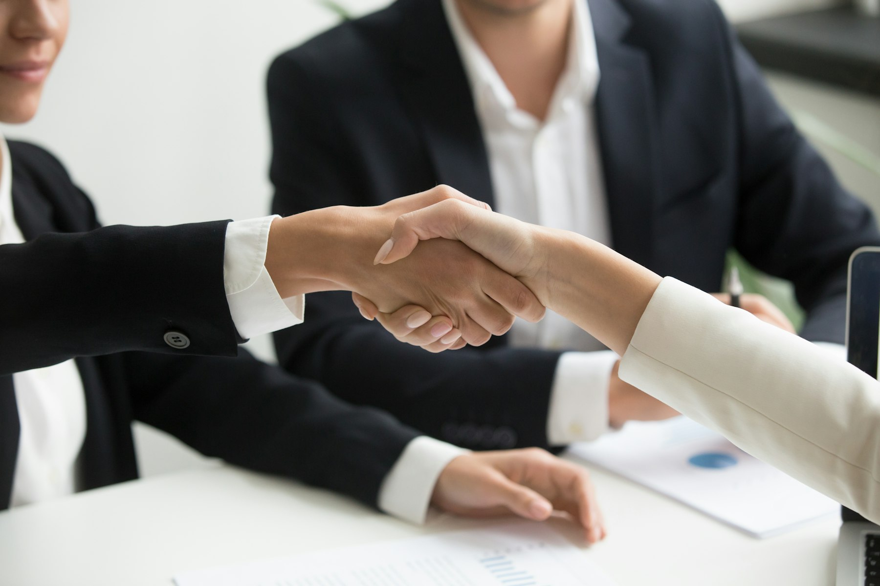 Two people shaking hands over a business deal at a table, colleagues in the background